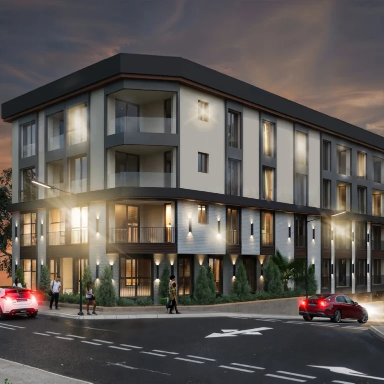 Bu görüntü, modern bir apartman binasını ve çevresini gösteriyor. Gece vakti çekilmiş olup, aydınlatılmış. Ön planda birkaç araç ve insanlar yürürken görünmekte. Arka planda ise ağaçlar ve bir manzara var. Bina, çağdaş mimarisiyle dikkat çekiyor ve sakin bir konumda yer alıyor.