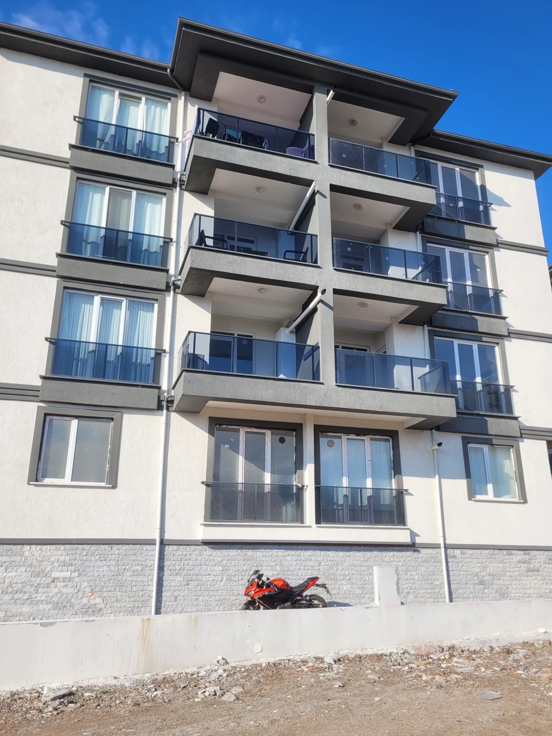 Troya Sitesi konut projesine ait dört katlı apartmanın balkonlu cephesi. Cam korkuluklu modern balkonlar ve binanın alt kısmında park edilmiş kırmızı bir motosiklet görülmektedir.