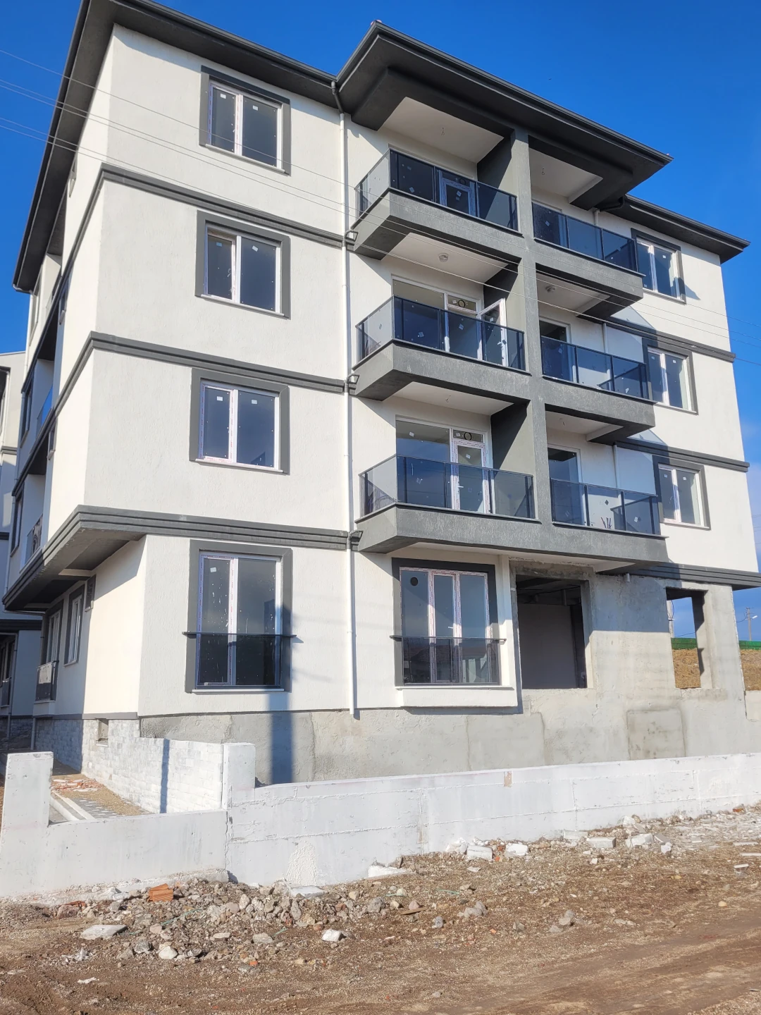 Yeni inşa edilmiş çok katlı bir apartmanın dış cephesi. Gri ve beyaz tonlarda modern tasarımlı yapı, cam korkuluklu geniş balkona sahip. Zemin kısmı henüz tamamlanma aşamasında; ön tarafta inşaat malzemeleri ve molozlar görülüyor. Gökyüzü açık ve güneşli.