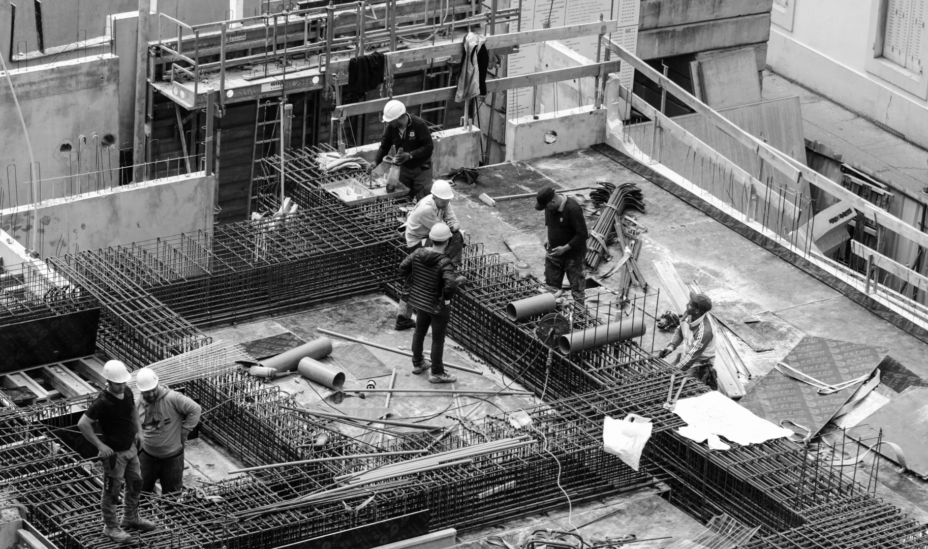 Bir inşaat alanında, işçilerin çeşitli görevleri yerine getirdiği bir sahne. İşçiler, beton dökme ve demir yapı üzerine çalışıyor. Bazı işçiler, koruyucu kask takarken, diğerleri el aletleri kullanıyor. Arka planda, inşaat malzemeleri ve yapının iskeletine ait demir ağları görünmekte. Alanın yoğun bir şekilde çalışıldığını gösteren bir atmosfer mevcut.